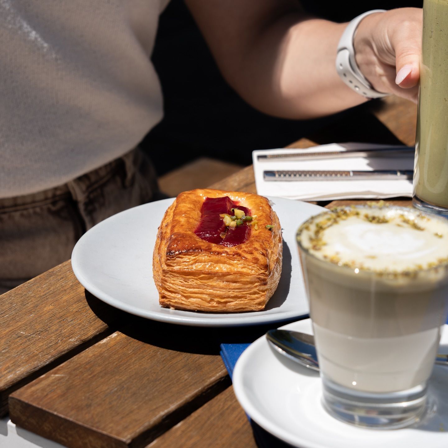 A jam-topped pastry in the sun beside a pistachio latte and a green smoothie on an outdoor table