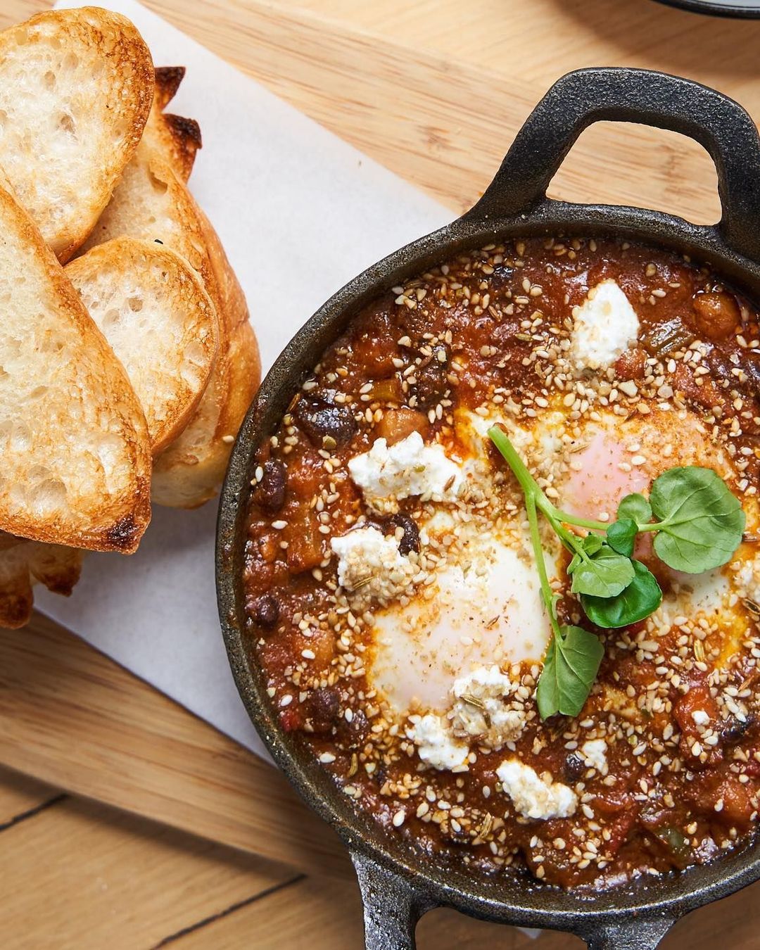 Baked eggs with tomato, chickpeas and feta in a cast-iron pan beside toasted sourdough