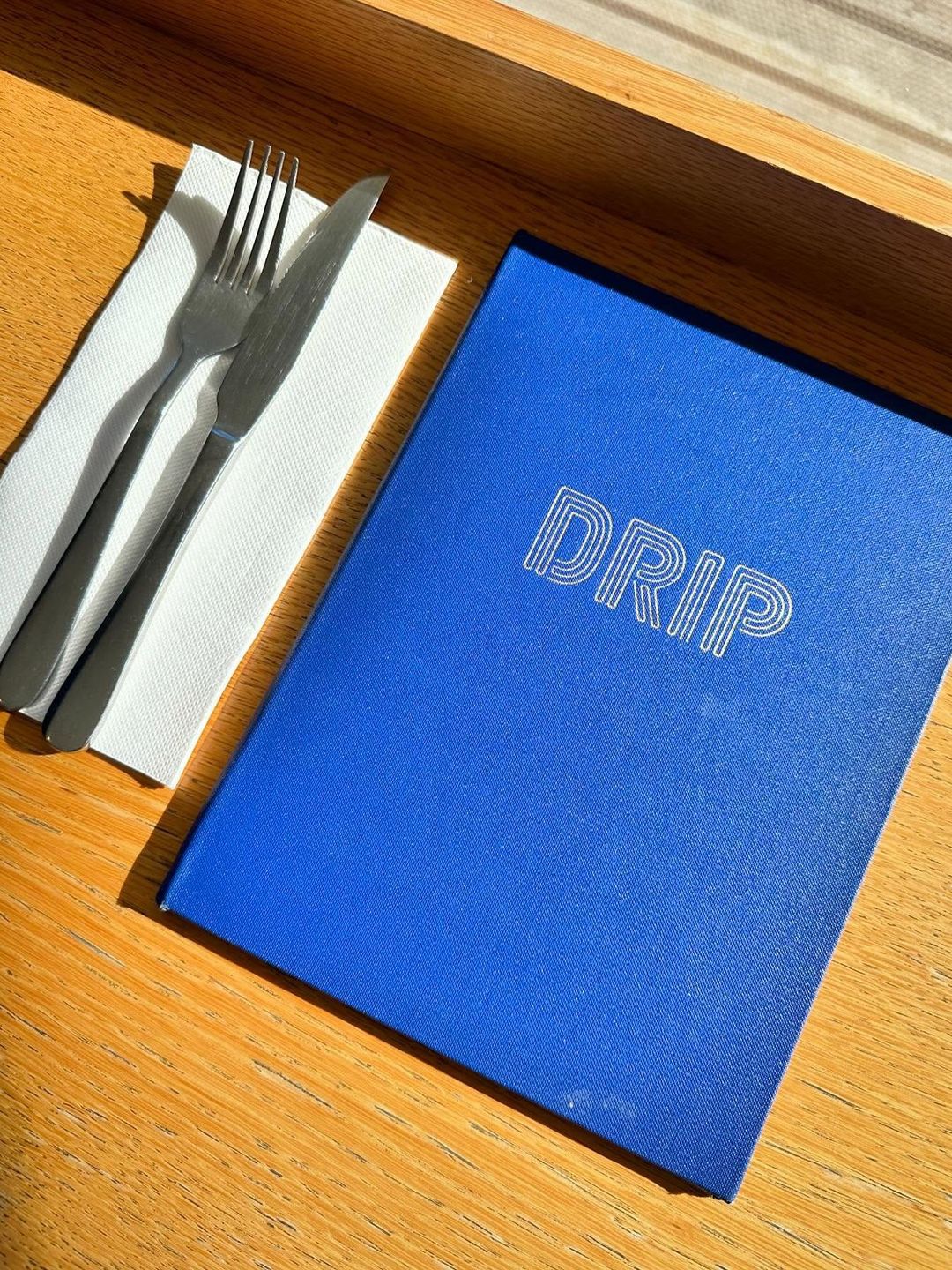 A blue DRIP menu and cutlery laid out on a sunlit timber table