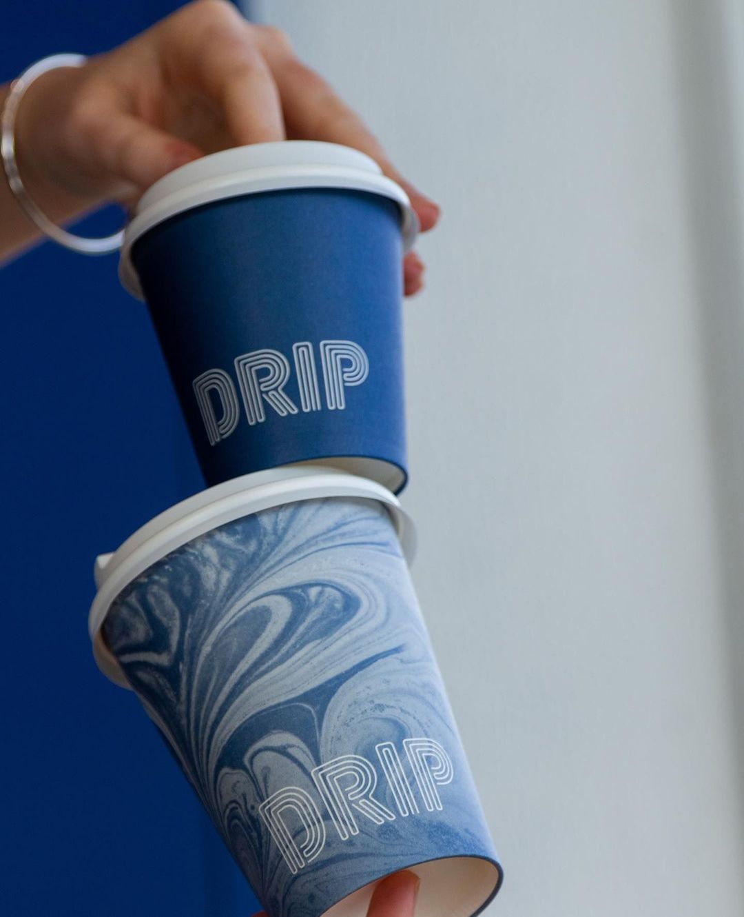 Two DRIP-branded takeaway coffee cups stacked and held against a blue wall