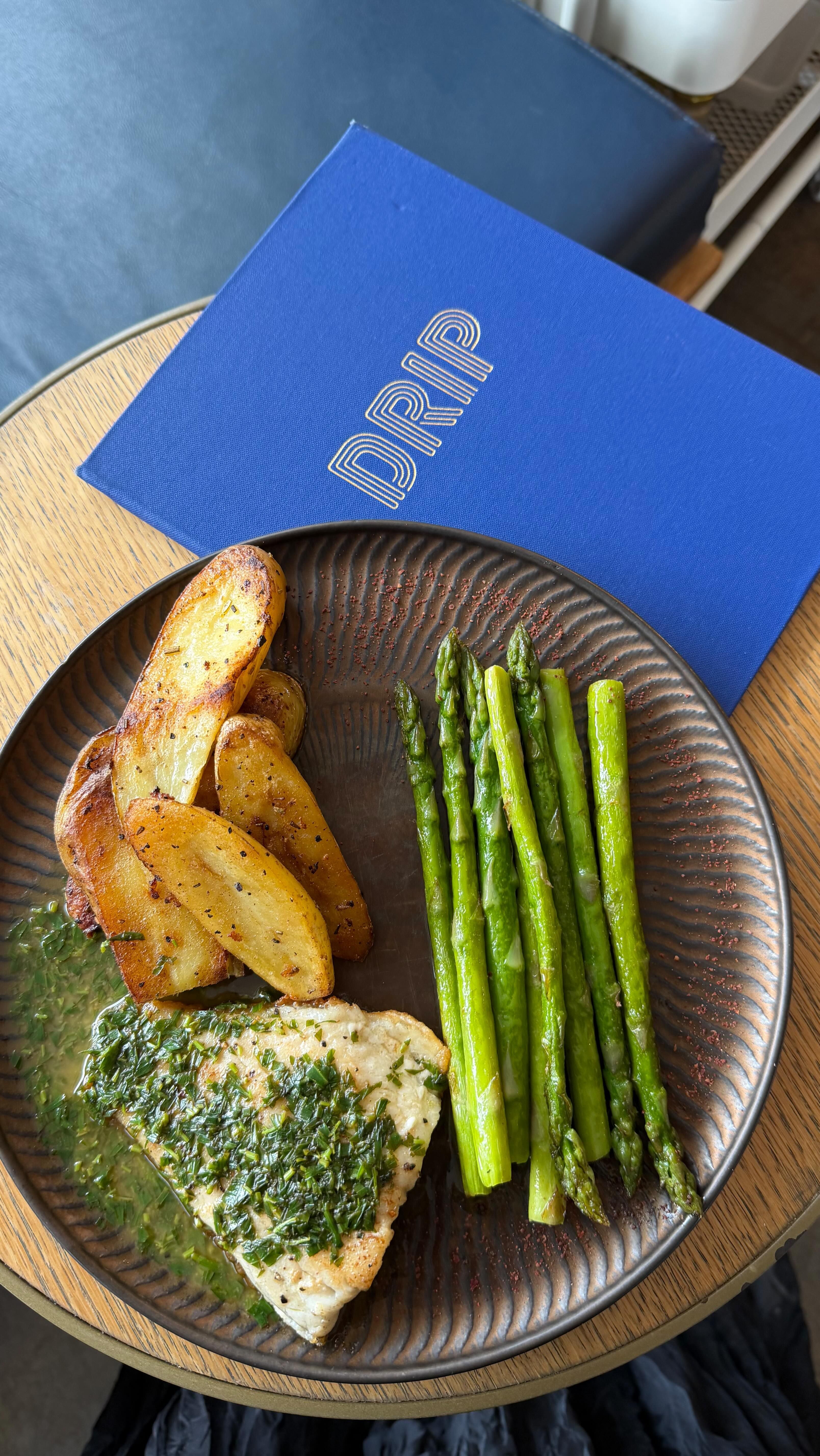 A dinner plate of fish with herb sauce, roast potato wedges and asparagus beside a blue Drip menu