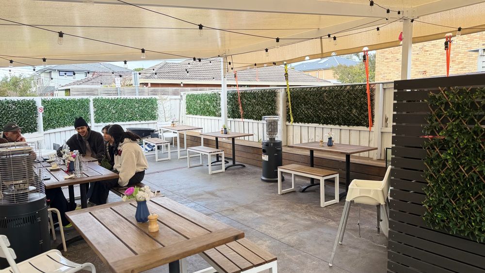 The covered courtyard seen from the far end, with timber tables and benches under a shade sail and festoon lights